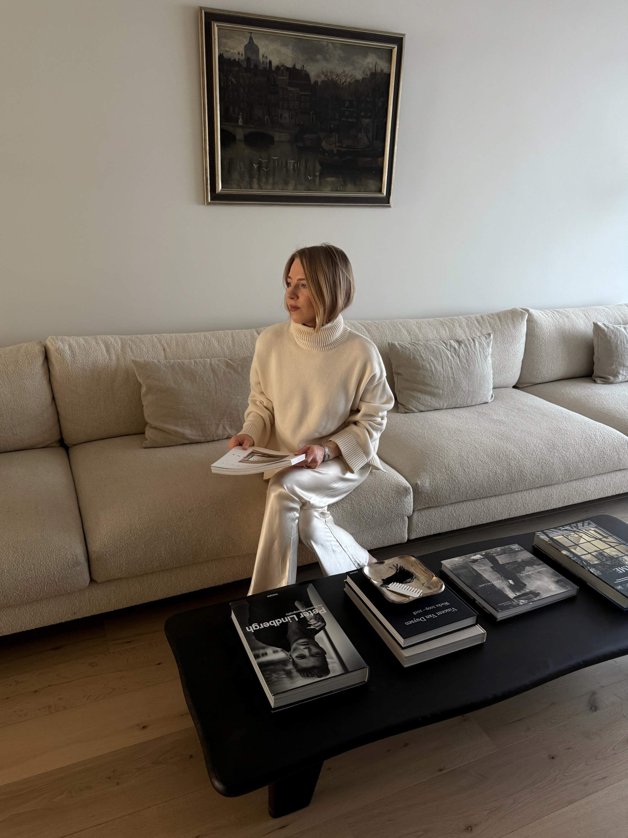 Creative Director Darya Burtseva seated on cream bouclé sofa reviewing design magazine, with vintage Amsterdam cityscape painting and black coffee table with art books