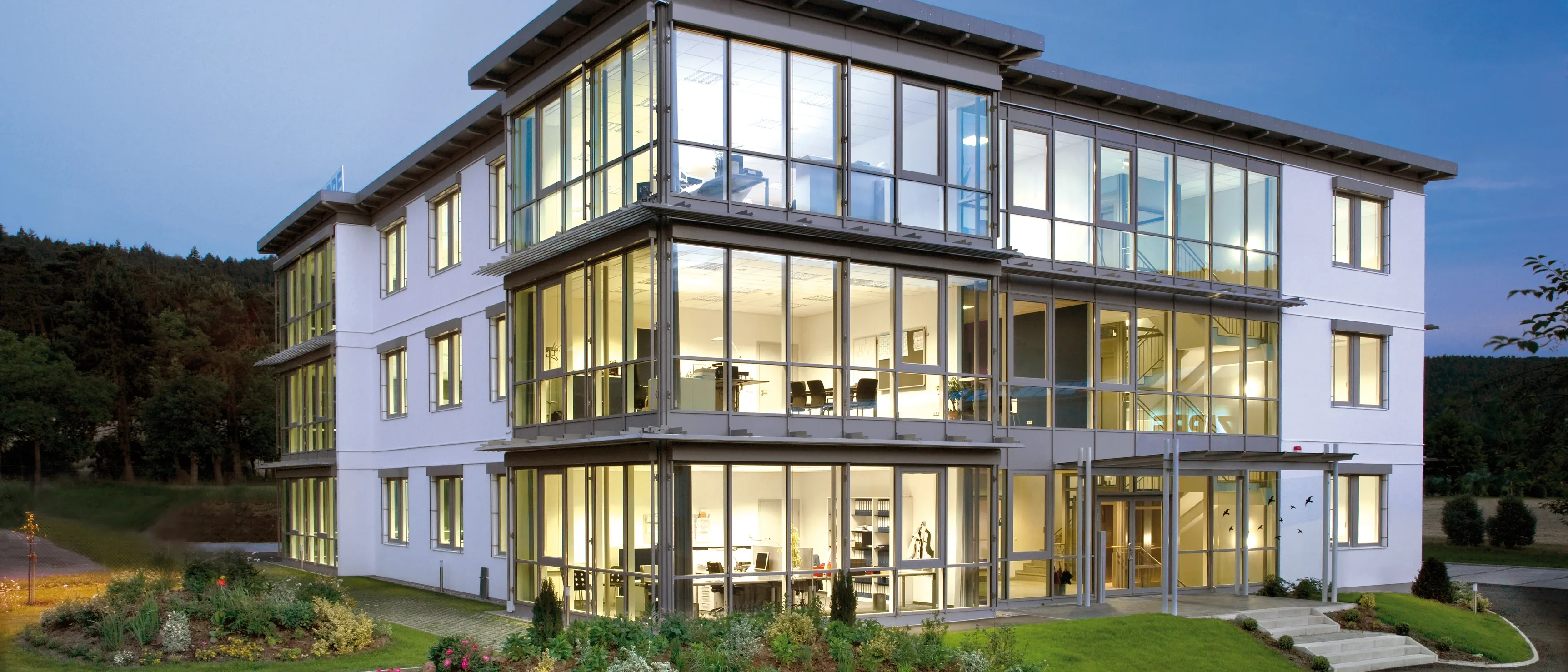 Modernes Bürogebäude mit viel Glas und Fassadenelementen in einer baumbestandenen Umgebung bei Dämmerung.