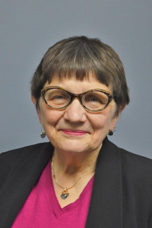 Portrait of an older woman with glasses, a pink shirt, and a black blazer, smiling at the camera.