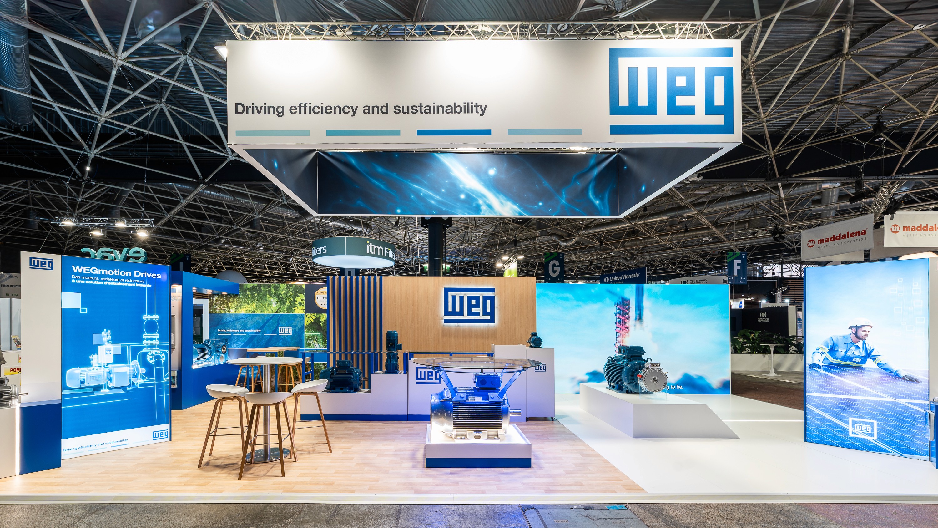 Stand WEG Pollutec 2025 réalisé par Expace – vue frontale du stand avec bandeau lumineux Driving efficiency and sustainability, moteur industriel sur podium vitré rétroéclairé, grands écrans LED et espace lounge bois