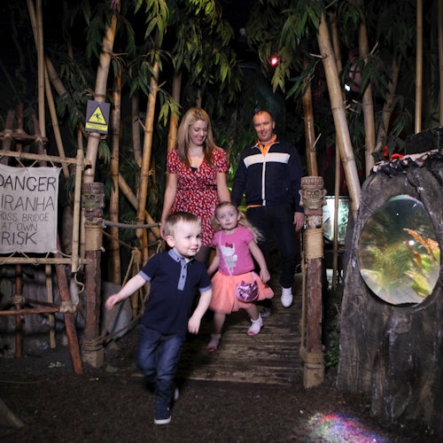 Two children running ahead of two adults on a wooden bridge in a jungle-themed setting with a sign warning about piranhas.