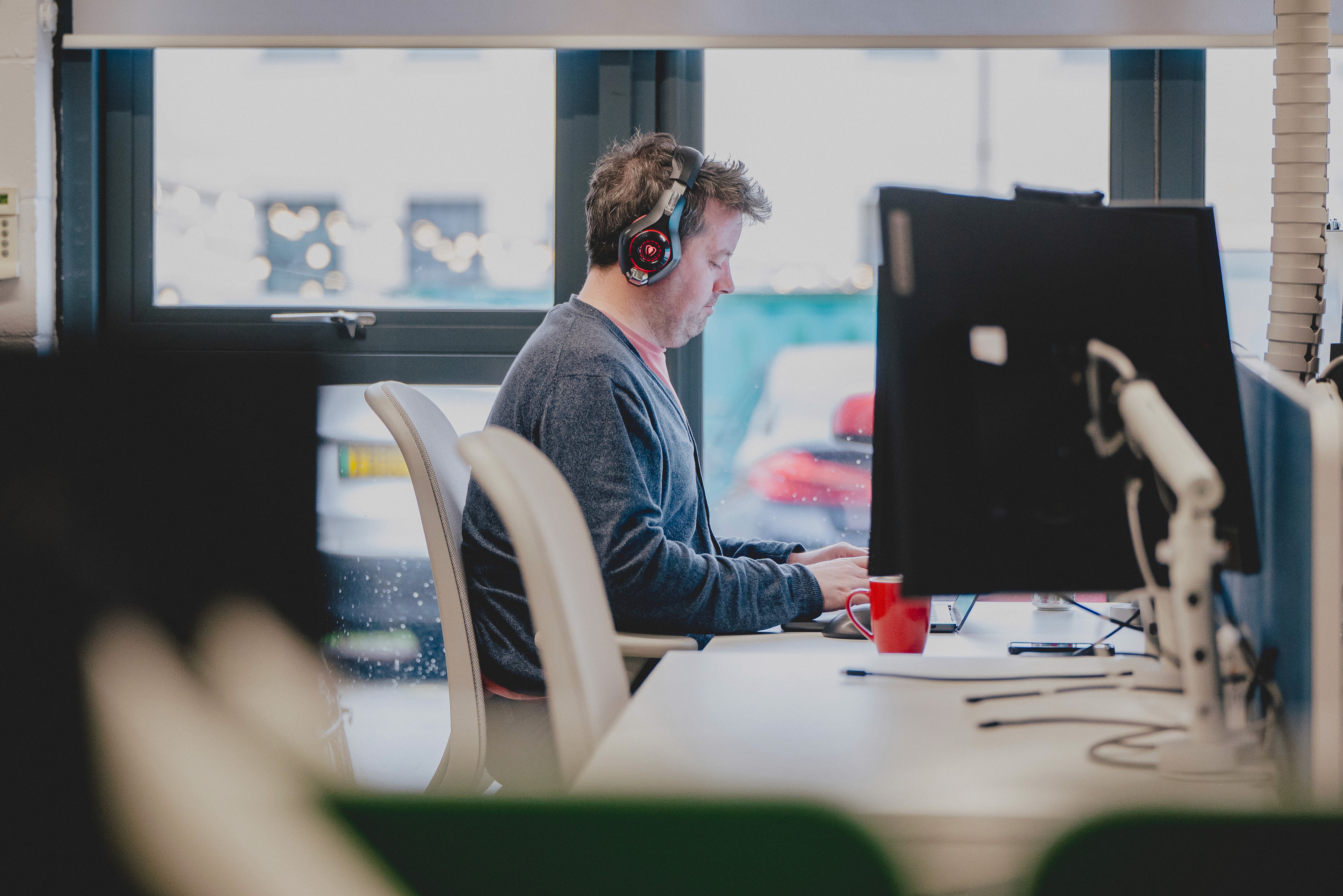 Person wearing headphones works at a computer desk in a modern office, with large windows and multiple monitors visible around the workspace.