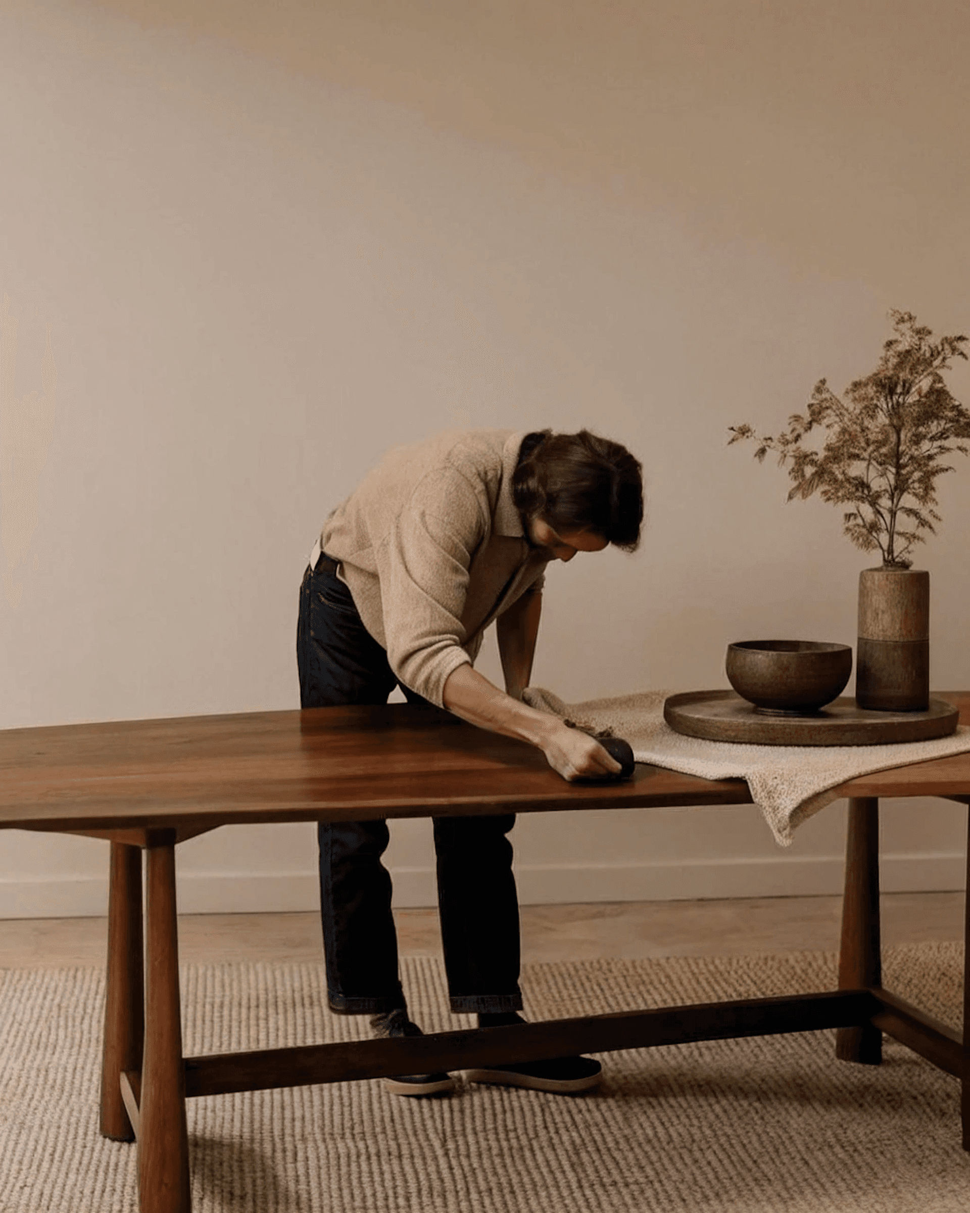 A person cleaning a polished wooden table in a minimalist room with neutral tones, featuring a vase, wooden bowl, and a textured table runner.