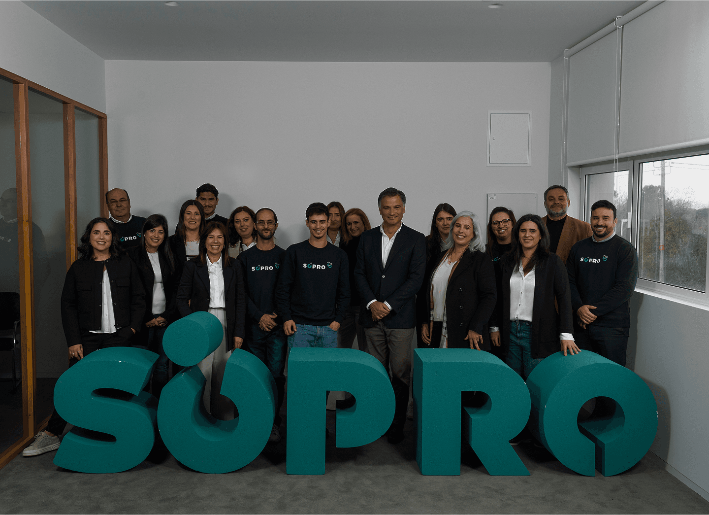 Grupo de pessoas sorrindo ao lado de grandes letras 3D formando a palavra  SRO, em um ambiente corporativo moderno, destacando a equipe da S/PRO na campanha de destaque na área de tecnologia e inovação.