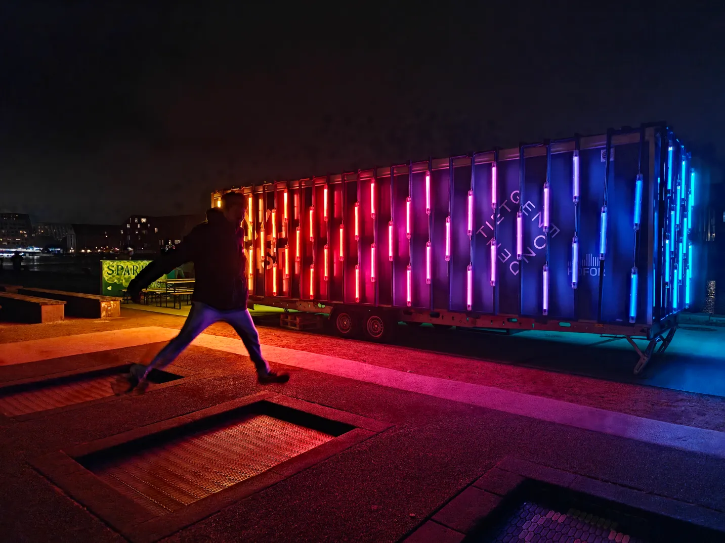 A person up in the air after jumping on a street trampoline with a colourful neon-light exhibition surrounded by darkness.