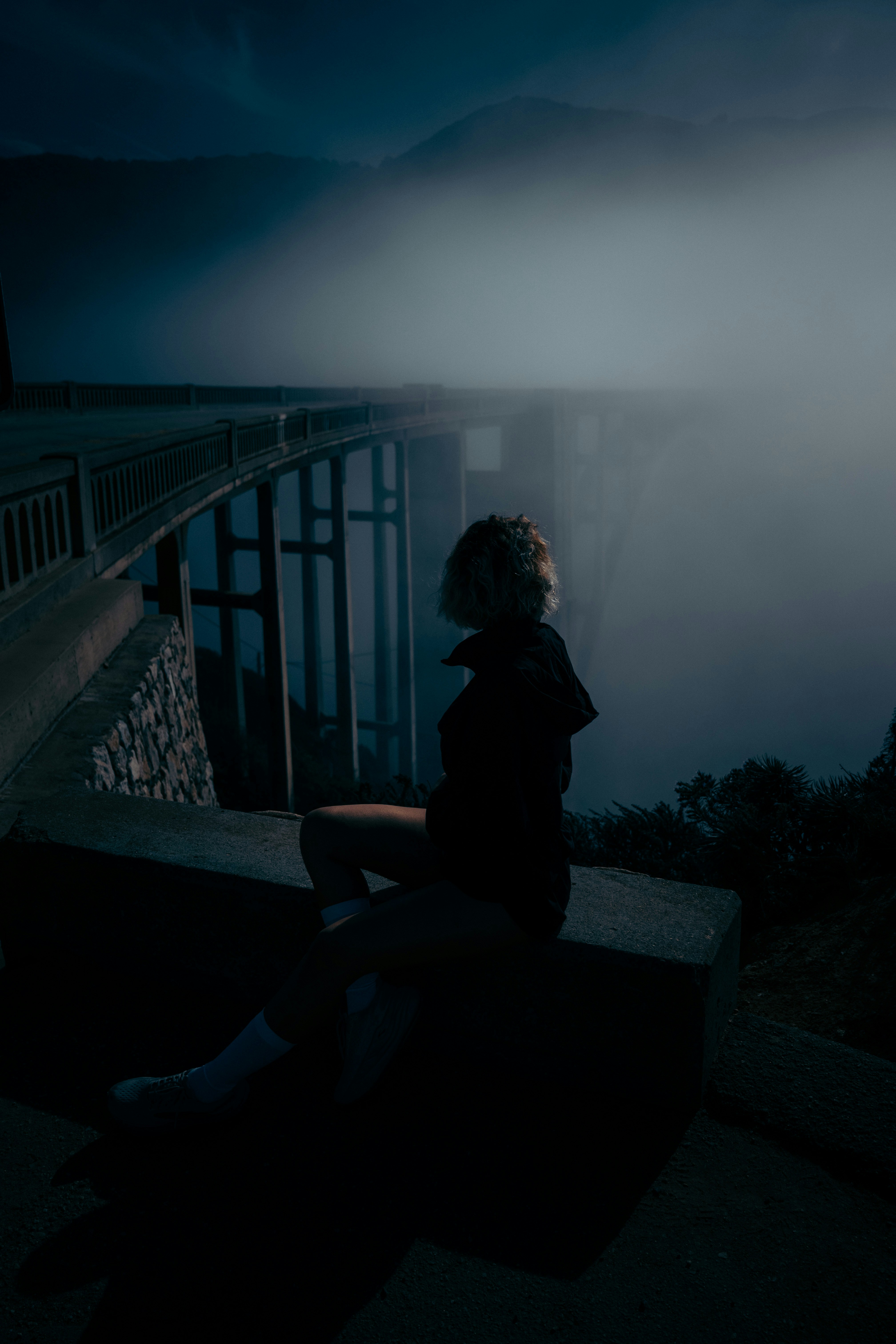 Woman sitting on ledge near foggy bridge at night