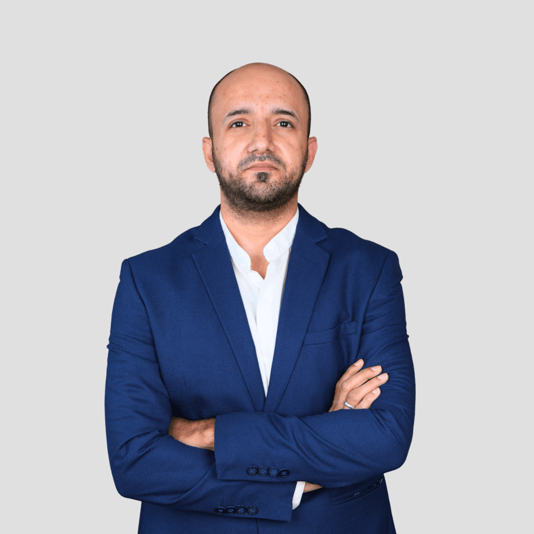 A woman sits at a desk with a laptop, laughing. A man stands beside her, pointing at the screen. The mood is collaborative and cheerful.