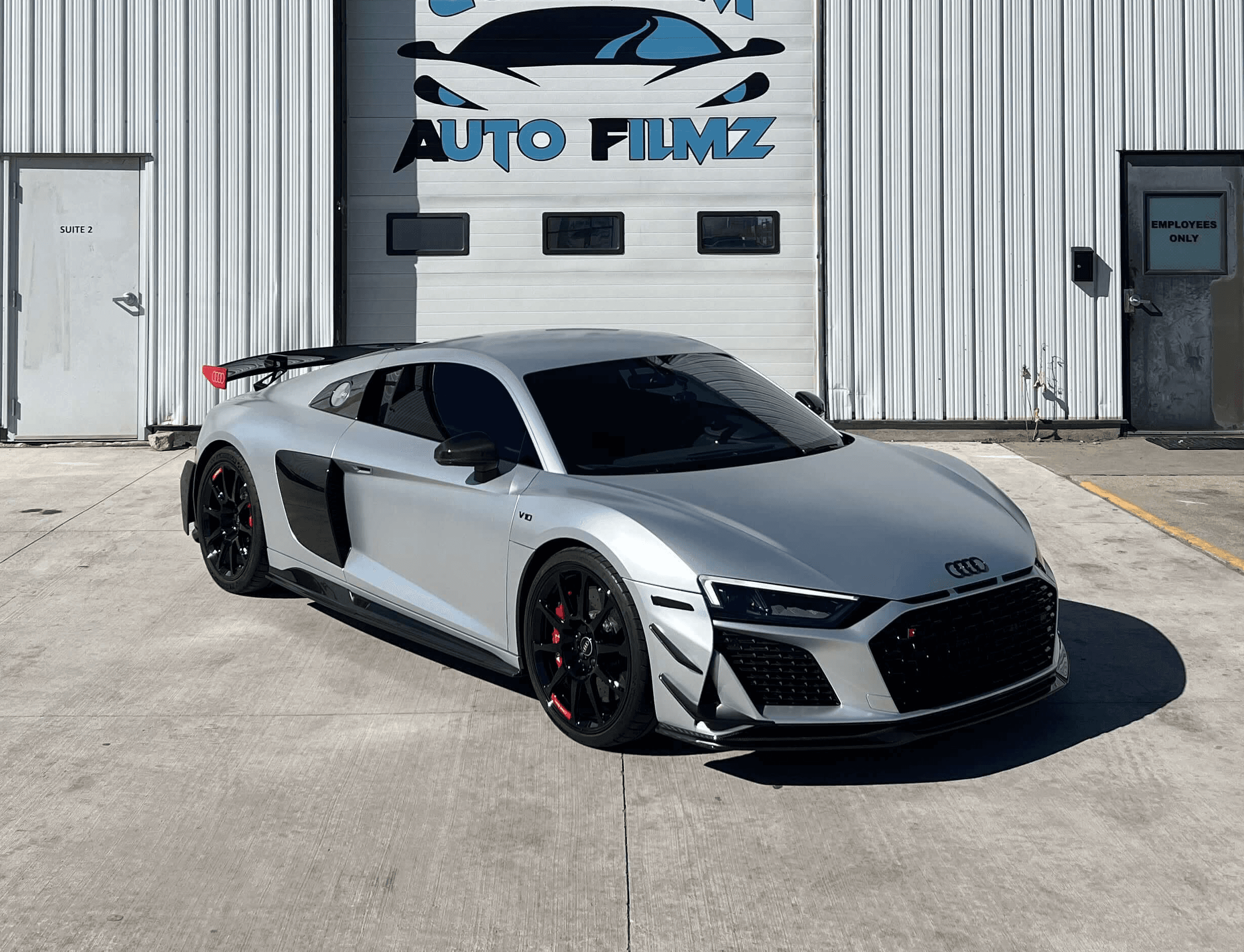 A sleek silver Audi R8 sports car is parked in front of a white industrial building with the sign "Custom Auto Filmz" displayed prominently above one of the garage doors.