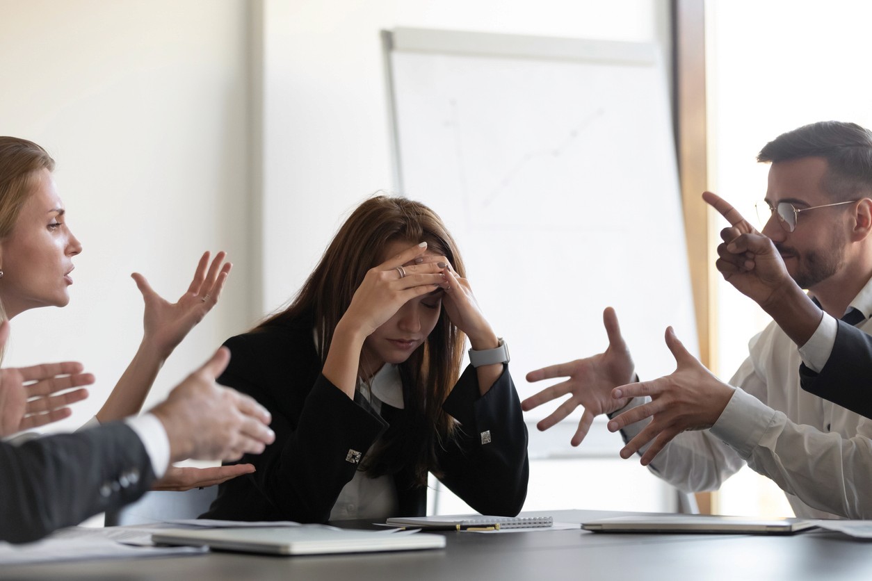 A woman sitting at a meeting table looking overwhelmed while colleagues argue around her.