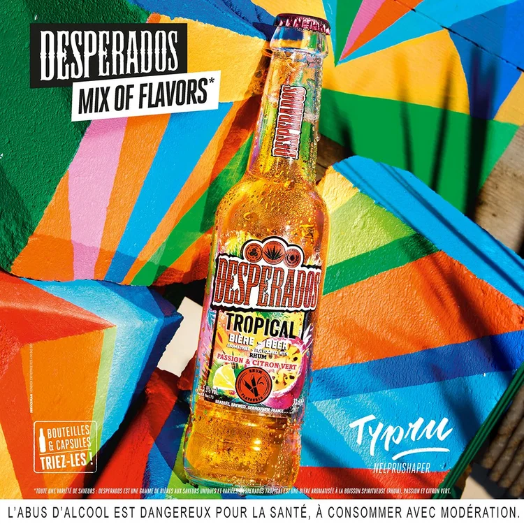 Close-up of a yellow Desperados tropical beer bottle with condensation, with a colorful, geometric background and a tote bag that says "TYLLU NEPRUSHAPER" in the background.