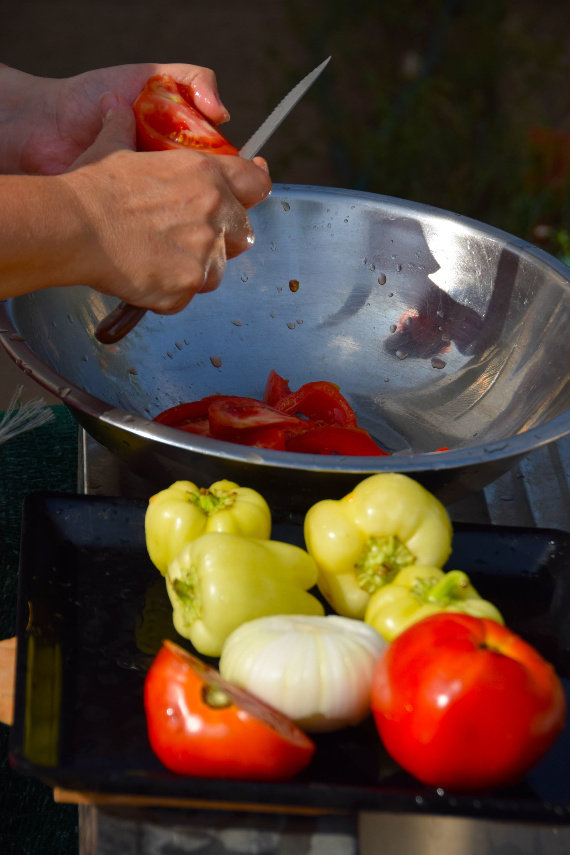 Person cutting tomatoes over a metal bowl with bell peppers and onions on a grill.