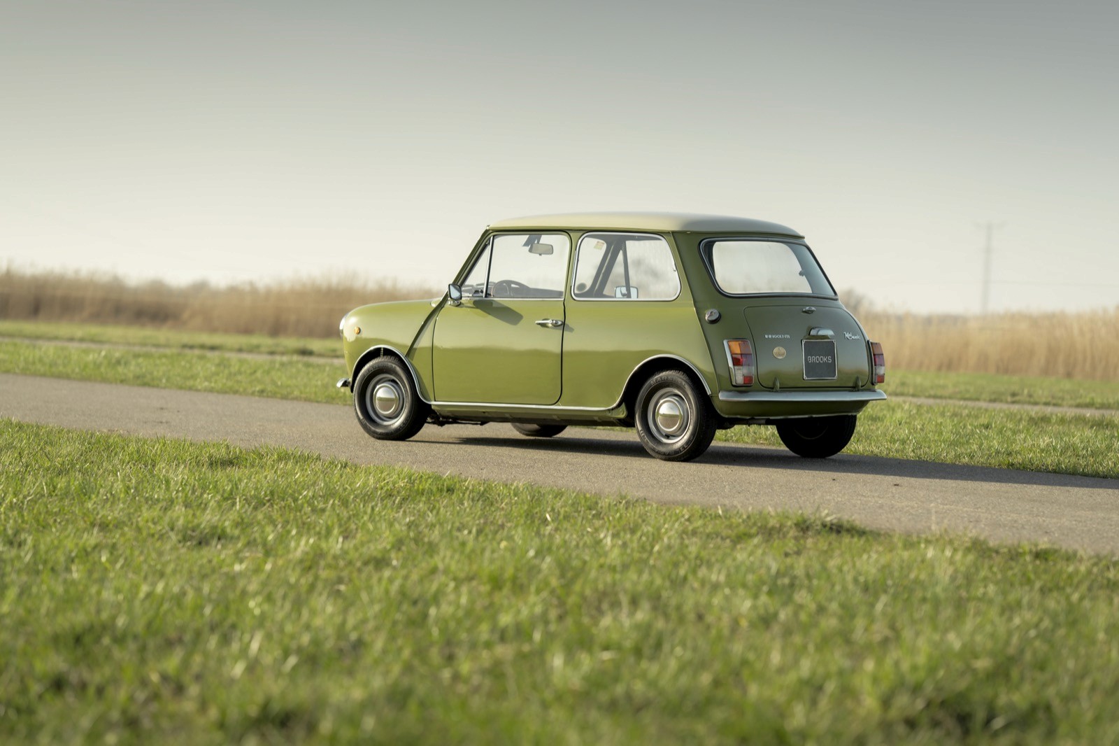 A classic green car drives on a narrow road surrounded by grassy fields under a clear sky.