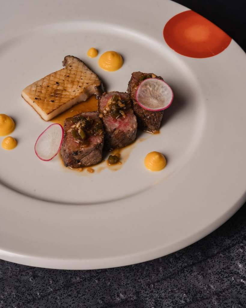 A beautifully presented dish featuring grilled beef with a garnish, accompanied by a slice of grilled fish and dollops of yellow sauce on a white plate.
