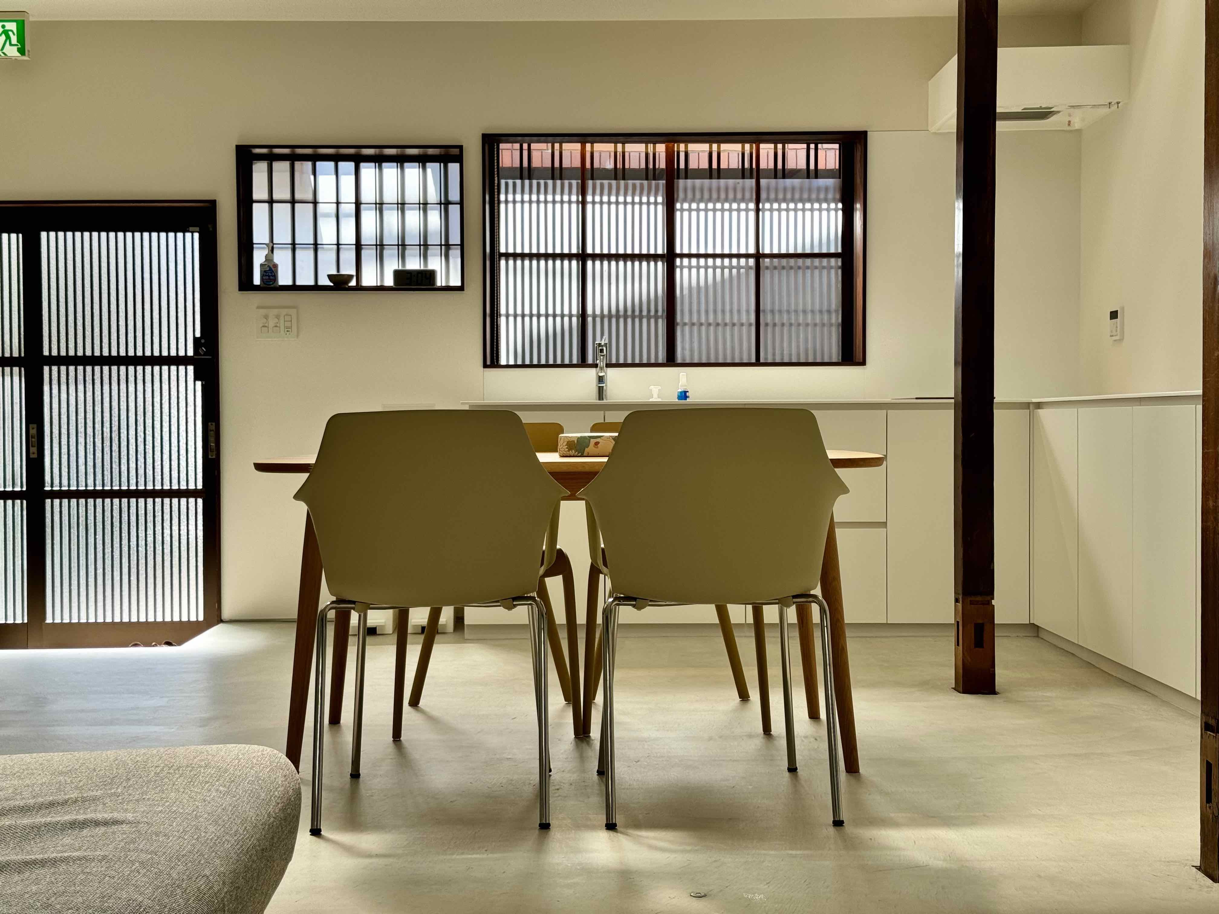 A minimalist dining room featuring a wooden table with two beige chairs, illuminated by natural light through large, grid-patterned windows, creating a serene and inviting atmosphere.