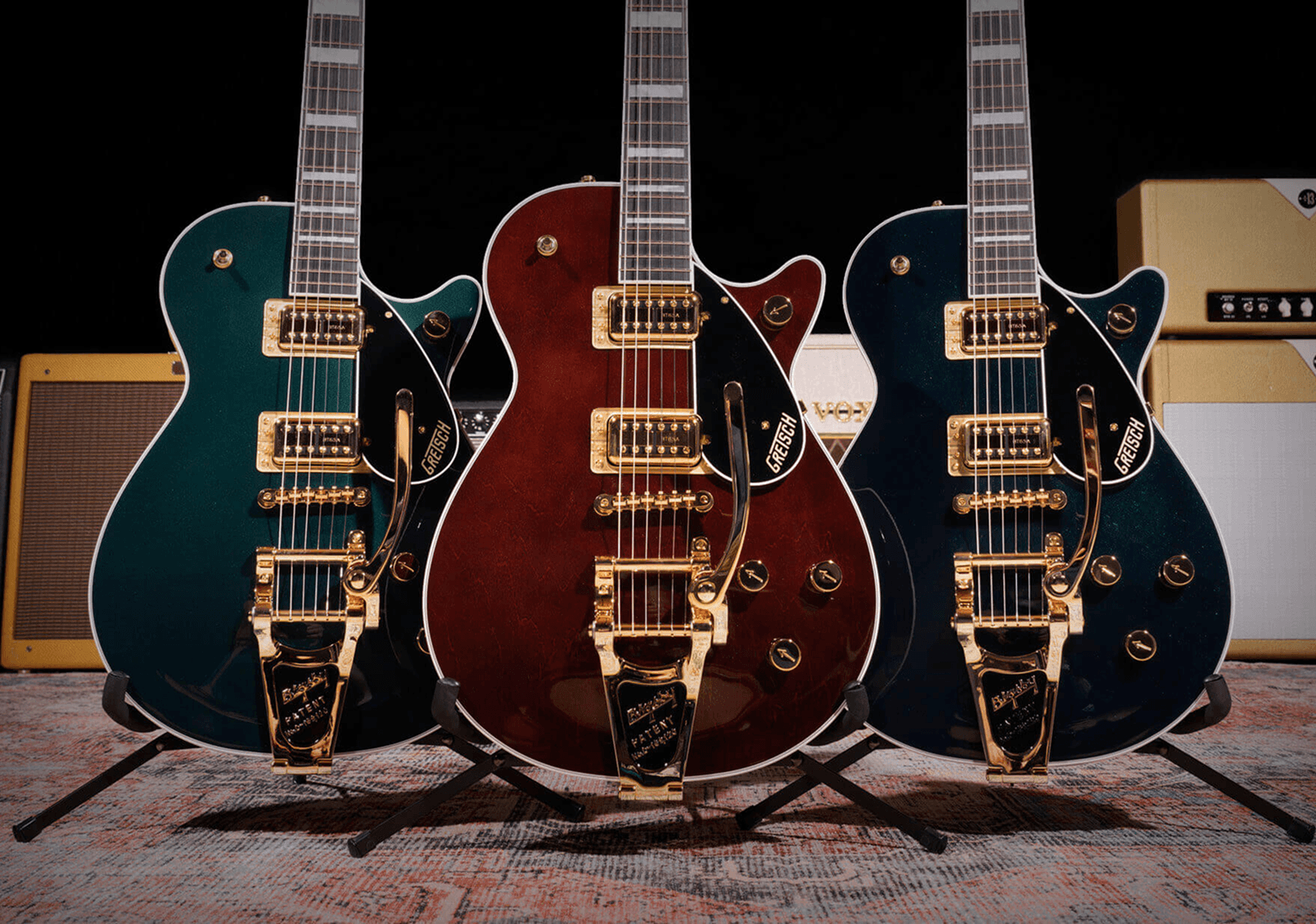 Three Gretsch solid body electric guitars in teal, burgundy, and navy with gold hardware, displayed on stands.