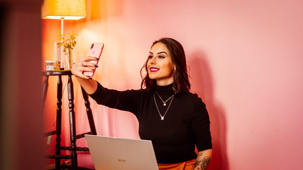 Influencer taking a selfie with her phone next to a laptop, illustrating Shopify Collabs