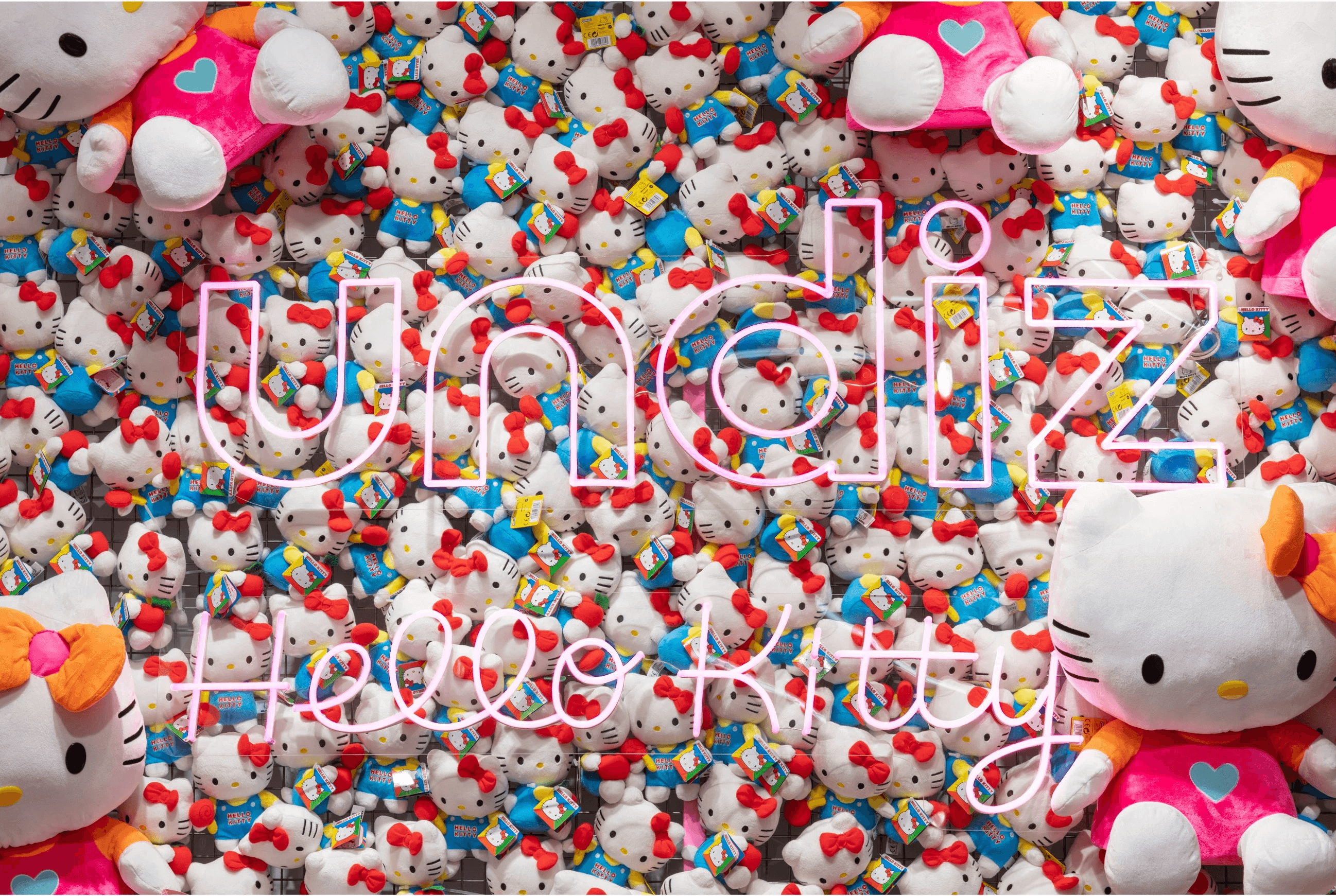 Enseigne lumineuse "undiz Hello Kitty" devant un mur recouvert de peluches Hello Kitty.