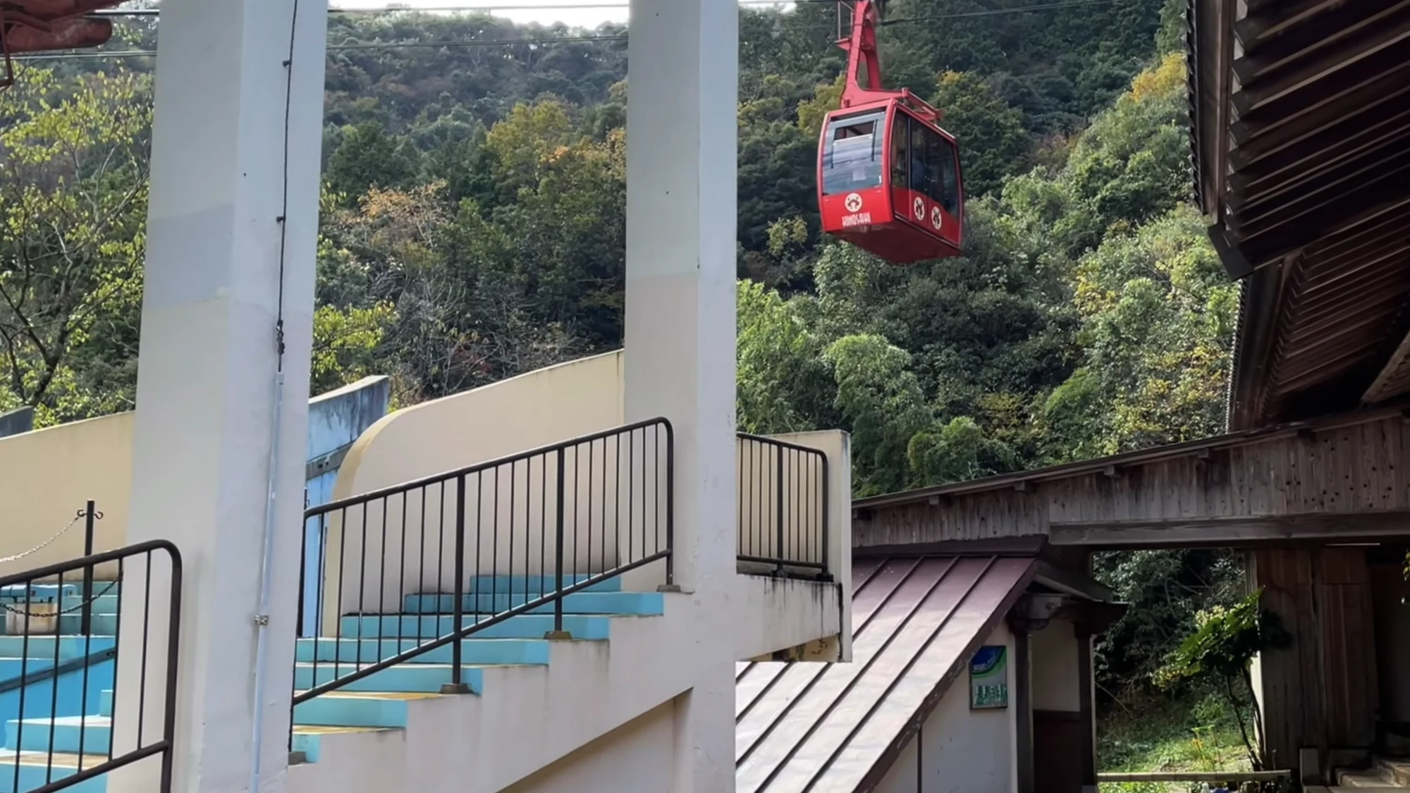 A red ropeway gondola seen from below climbing the wooded hillside above Kinosaki.
