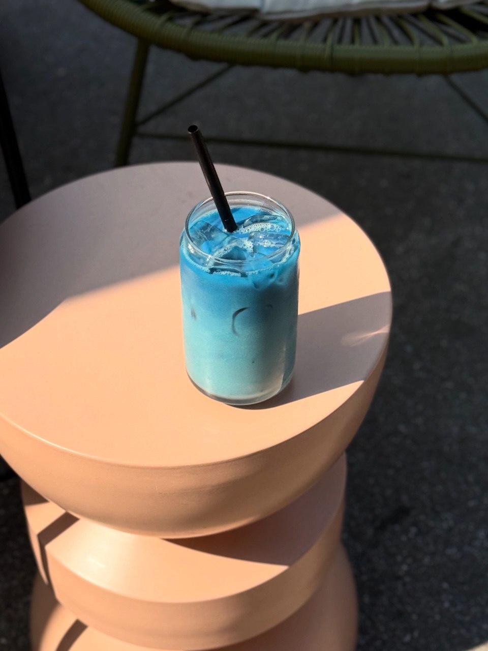 A refreshing blue iced drink in a clear glass with a black straw sits on a stack of round, peach-colored tables outside.