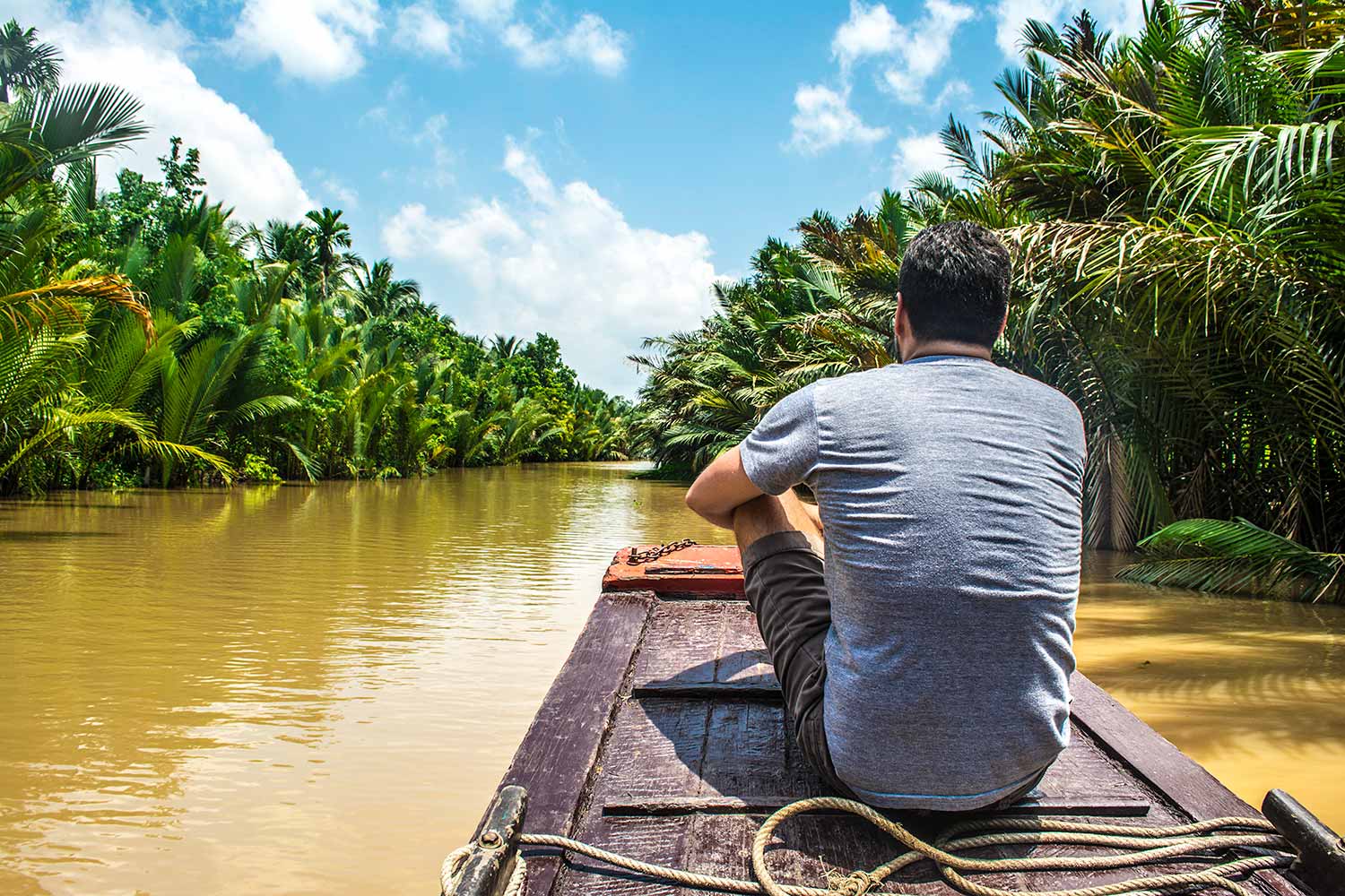 A man in a grey t-shirt sits on a wooden boat traveling down a muddy river lined with palm trees under a blue sky.
