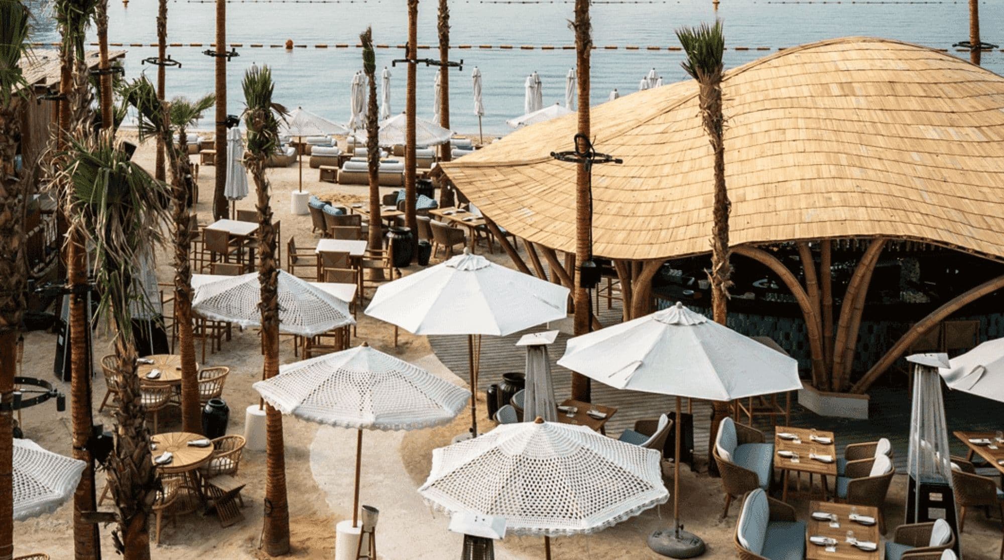 Koko Bay with a dining venue with a curved wooden roof, palm trees, white umbrellas, and outdoor dining areas.