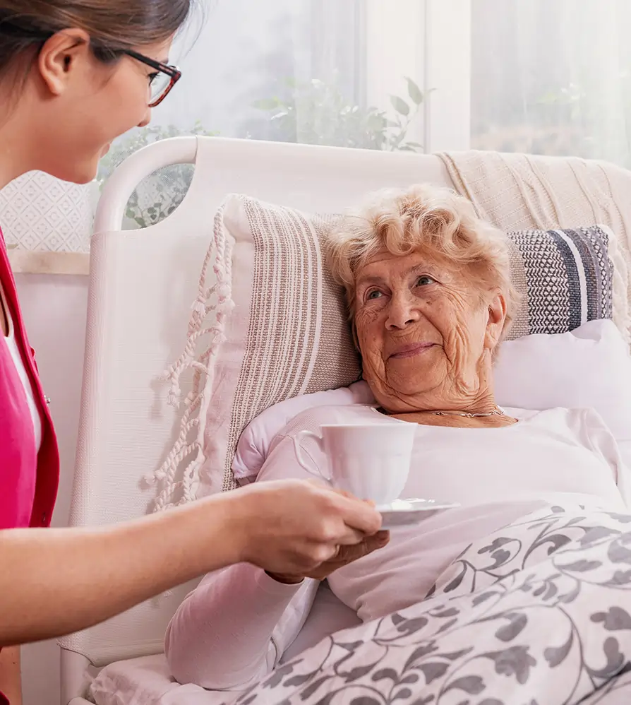 Caregiver offering a warm drink to a senior woman resting in bed during in-home hospice care