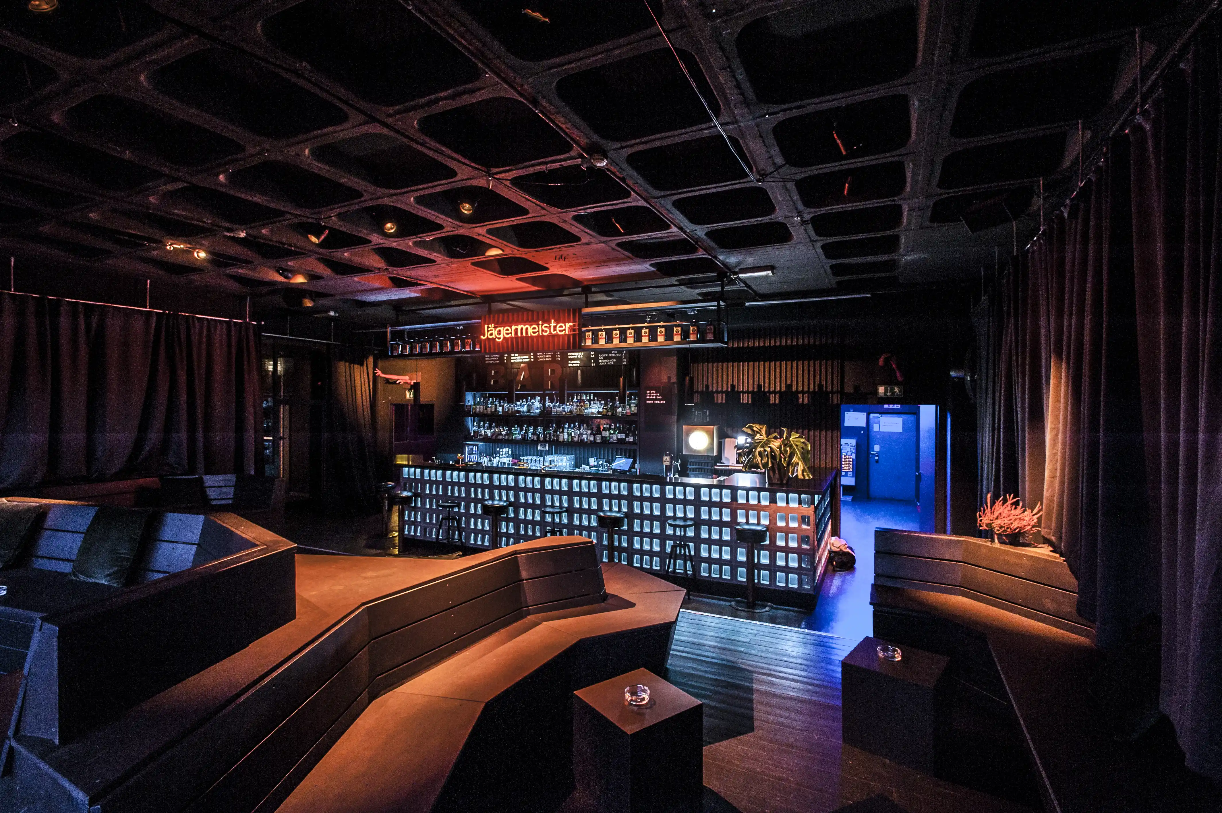 Dark club lounge of the Jägermeister Night Embassy with angular seating islands facing the backlit bar under a coffered ceiling.