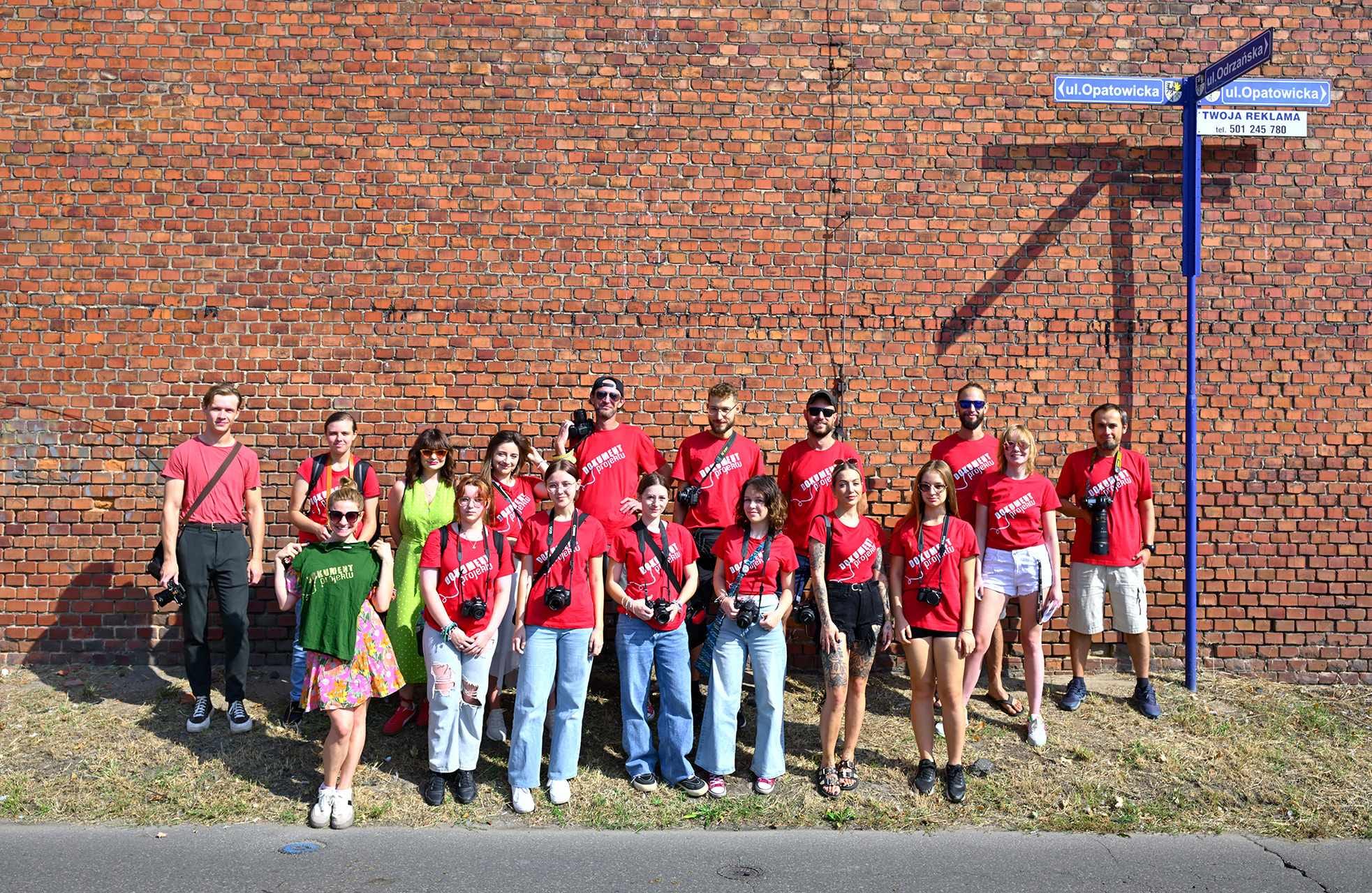 Grupa ludzi w czerwonych koszulkach stojąca obok znaku na tle ściany z czerwonej cegły