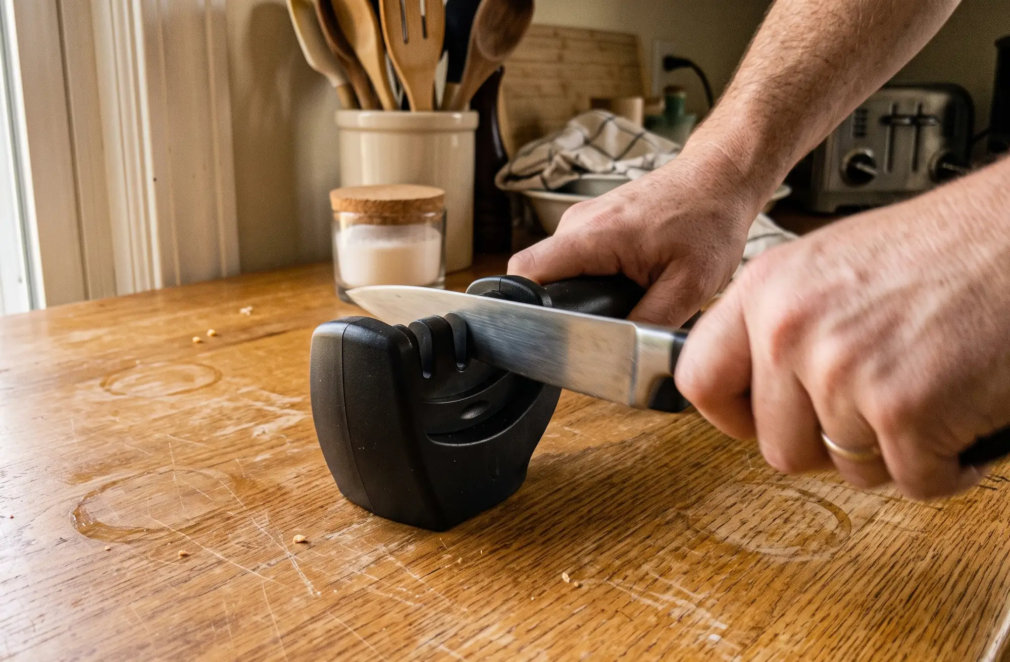 Person sharpening a kitchen knife by pulling it through a handheld black pull-through sharpener on a wooden table.