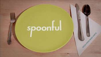 A light-colored wooden table holds a lime green plate with the word "spoonful" in white letters. To its left is a silver fork, and to its right are a silver knife and a black spoon. A white napkin with the words "LOCAL FOOD MARKET" lies beneath the utensils.