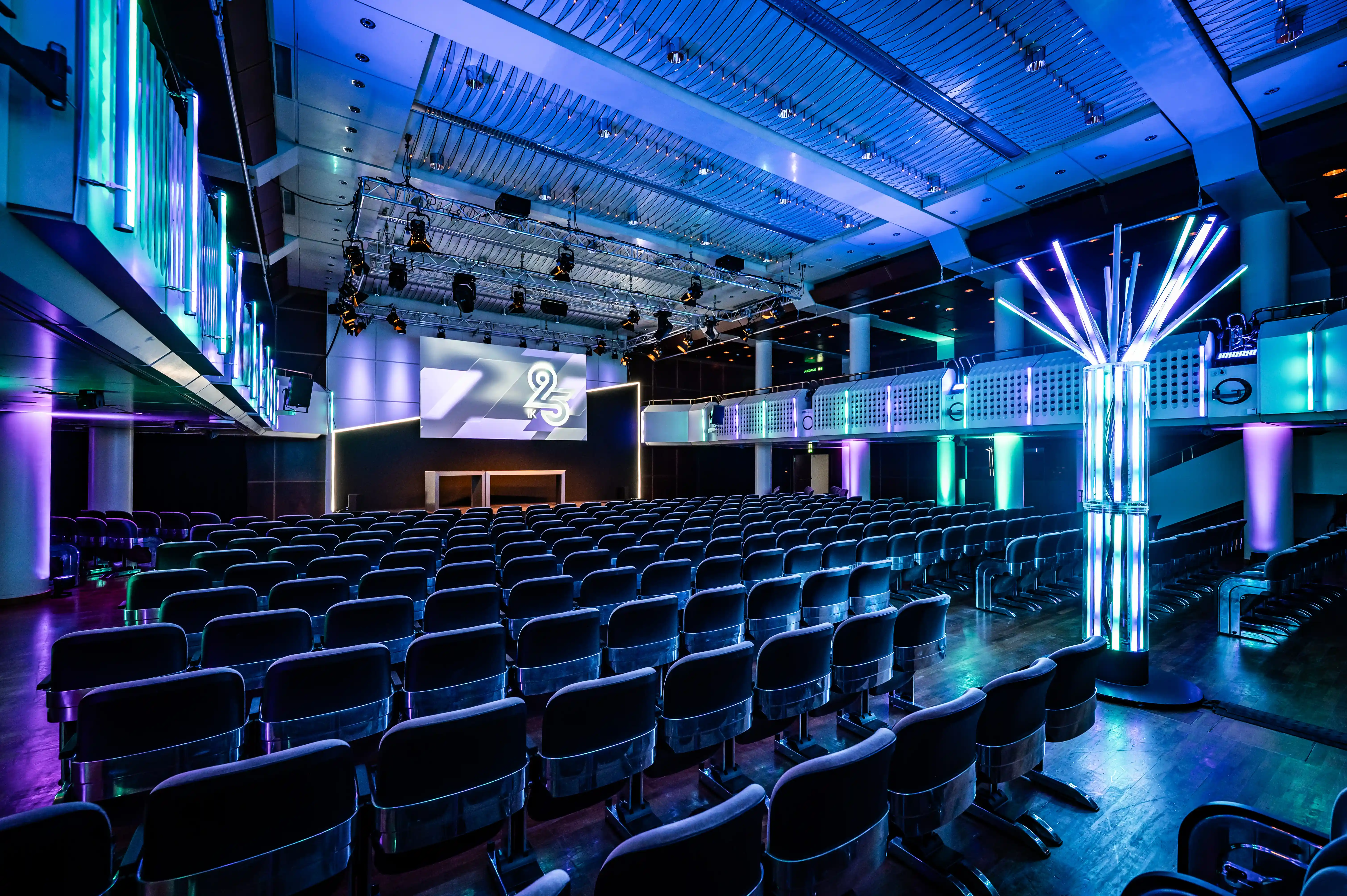 Blue-lit auditorium with rows of seats, glowing light tree pillar and 25 logo screen.