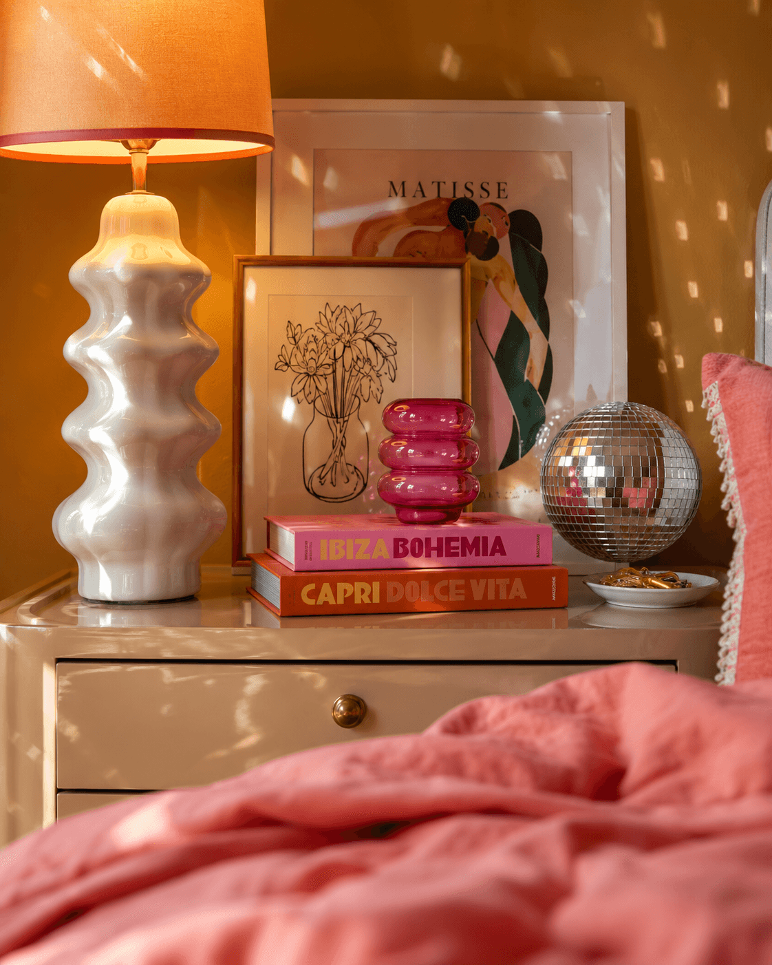 Modern dopamine decor bedside table vignette with sculptural ceramic lamp, mustard wall, coral bedding, colorful stacked design books, neon pink glass decor, layered framed artwork, and disco ball reflections in a playful yet sophisticated maximalist bedroom interior.