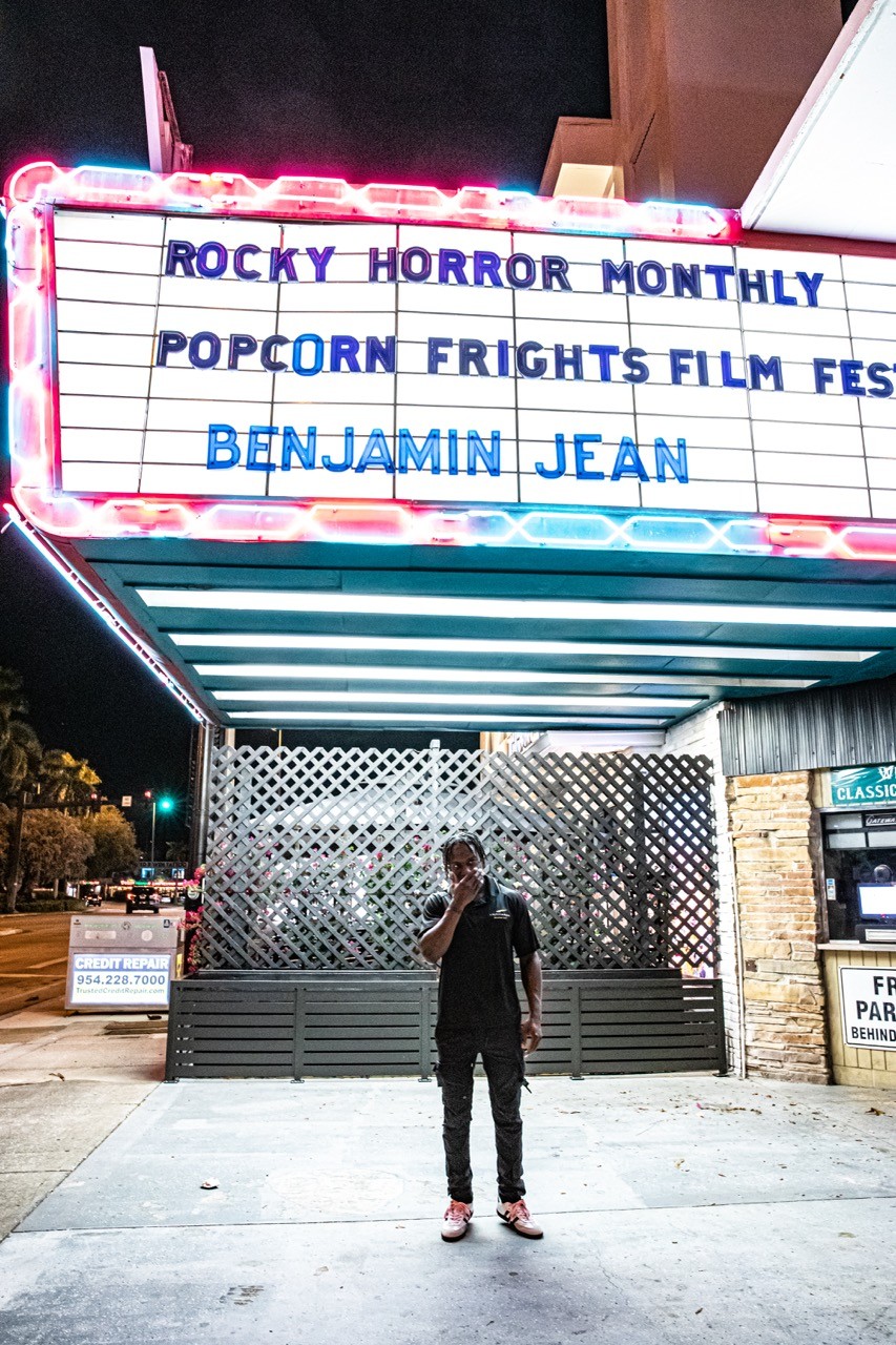 Night exterior of a theater marquee reading “Benjamin Jean,” with Benjamin standing on the sidewalk below.