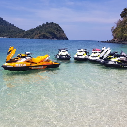 在藍天之下，海灘附近的清澈淺水中停著摩托艇，背景是一個有丘陵的小島。