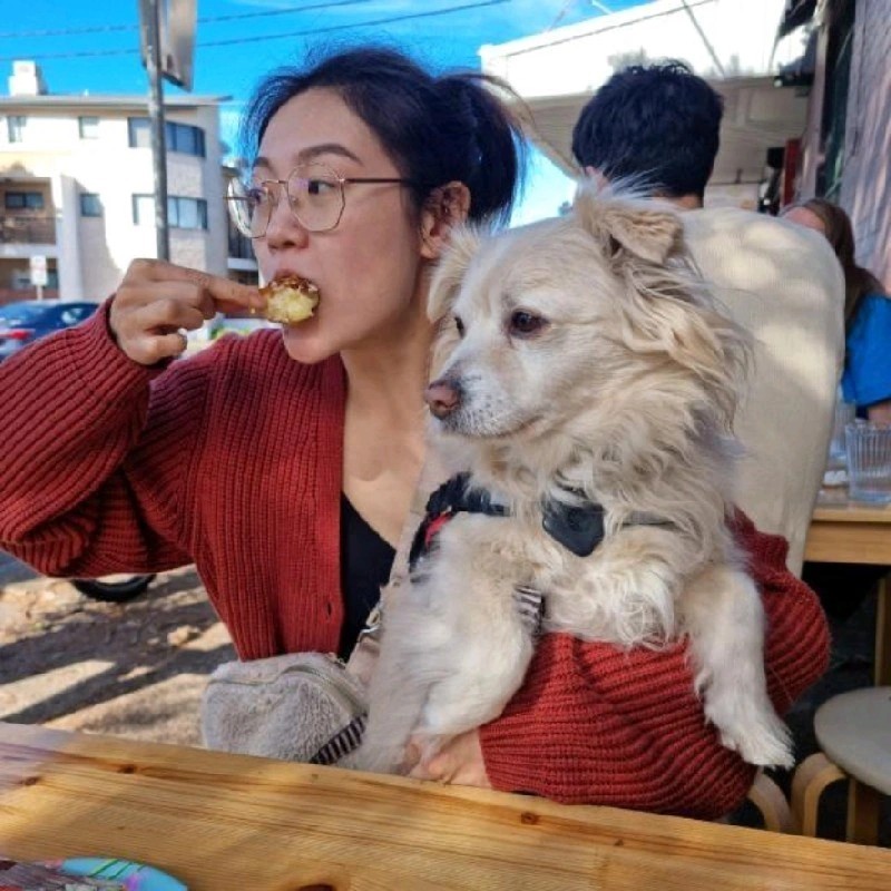 Woman wearing glasses and red cardigan eating while holding a golden retriever dog at an outdoor venue.