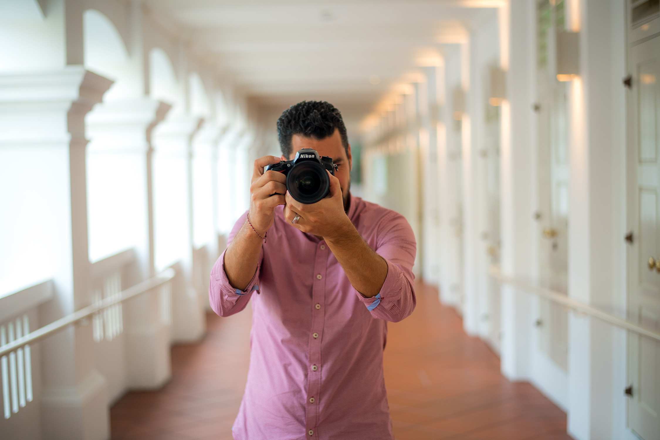 Man holding a camera stands in a bright, long hallway with white railings and warm wooden floors under soft natural light.
