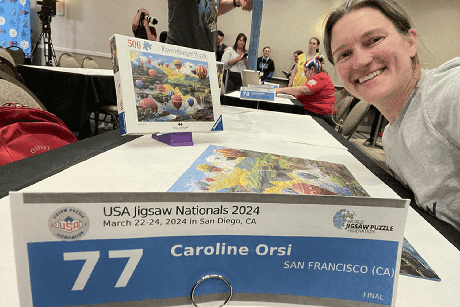 In a lively event hall, participants involved in a jigsaw puzzle competition work diligently at tables, with one table labeled "77" and a smiling woman posing nearby under a sign for "USA Jigsaw Nationals 2024" in San Diego, California.