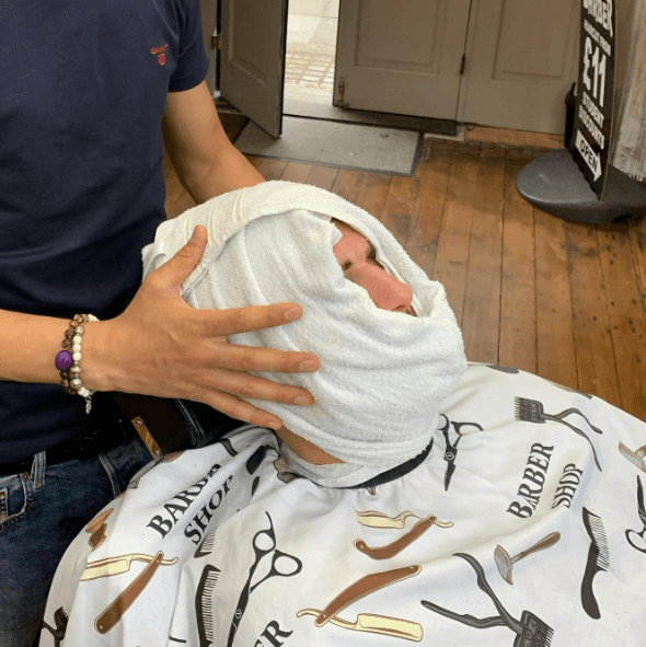 Man with a heated towel receiving a facial service, seated comfortably in a relaxed position.