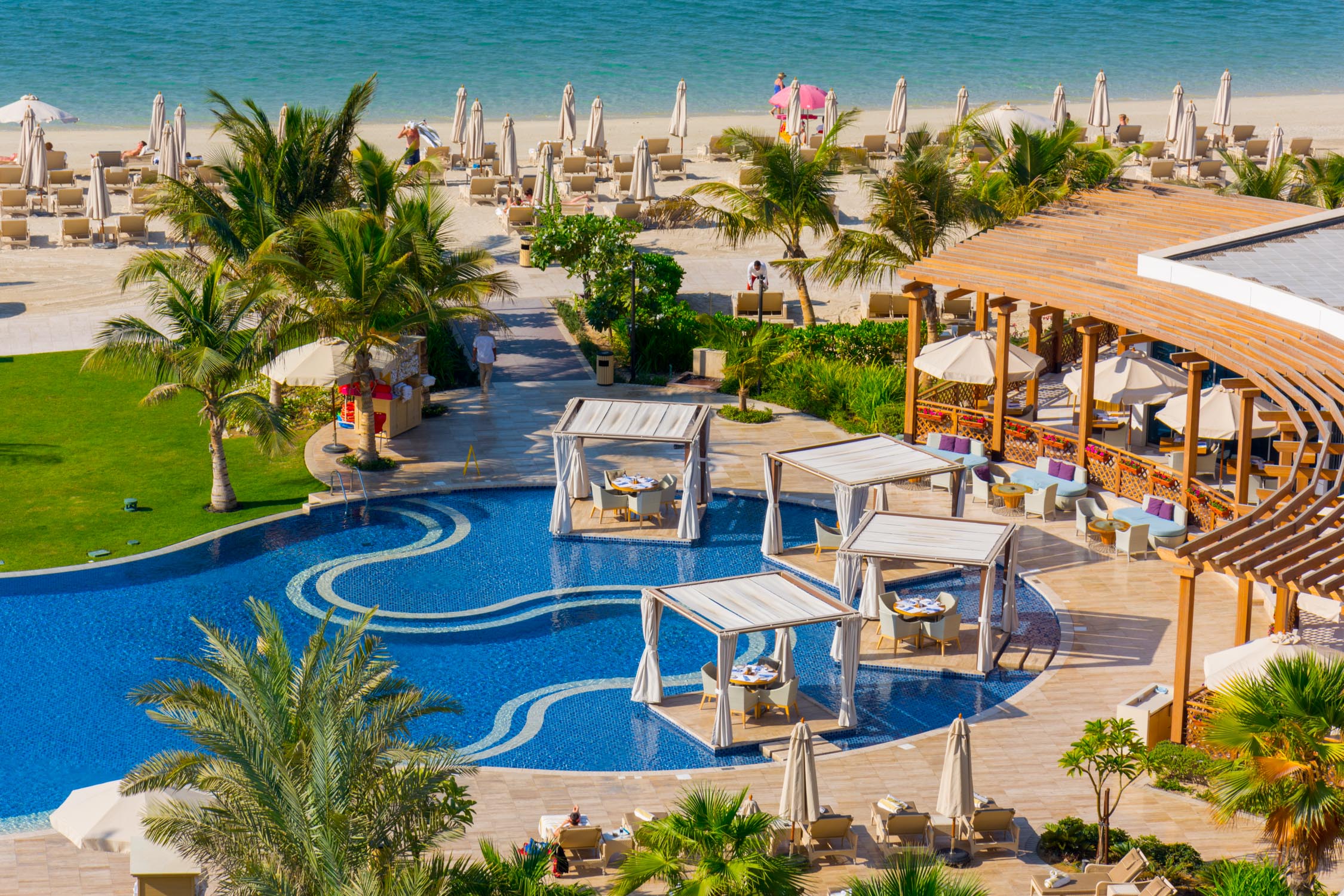 A tropical beach resort with a blue swimming pool featuring white cabanas and lounge chairs under palm trees by the sea.
