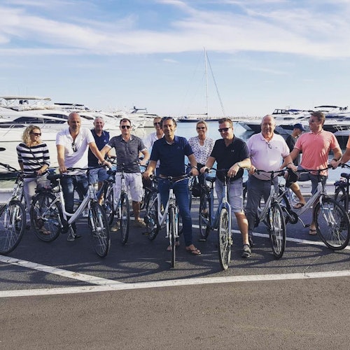 A group of nine people stand with bicycles on a waterfront, with boats and a clear blue sky in the background.