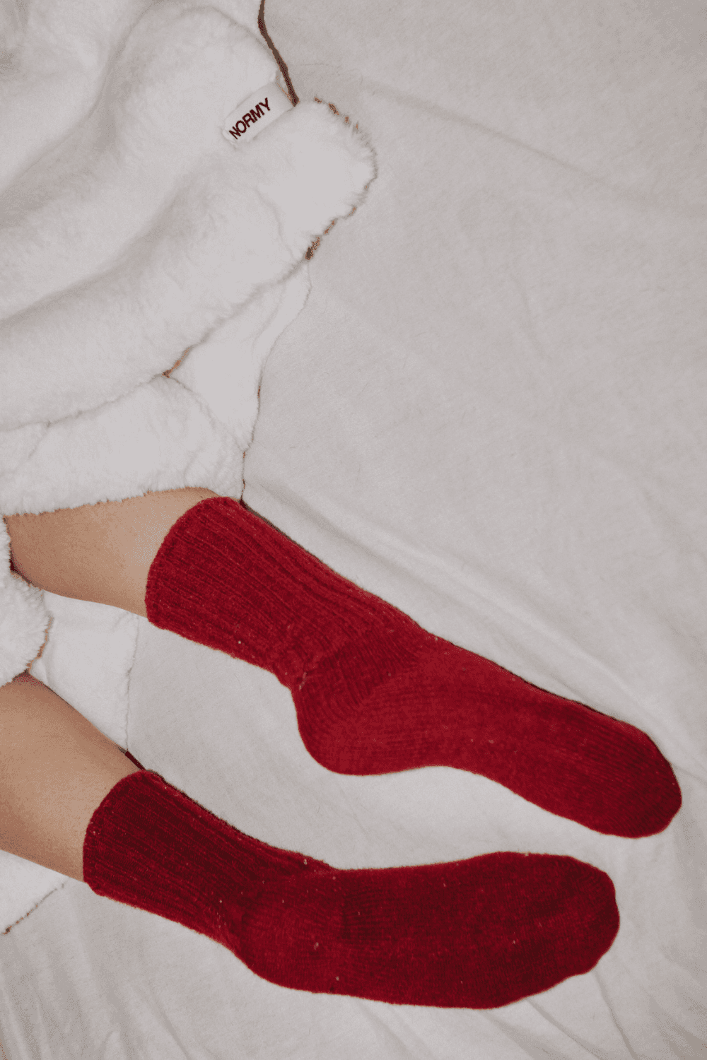 A cozy scene showing a person dressed in white lounging on a sofa, surrounded by plush blankets in cream and deep teal. Both blankets feature the "NORMY" label, highlighting the brand, and add a touch of comfort and style to the setting, which also features colorful tile flooring.