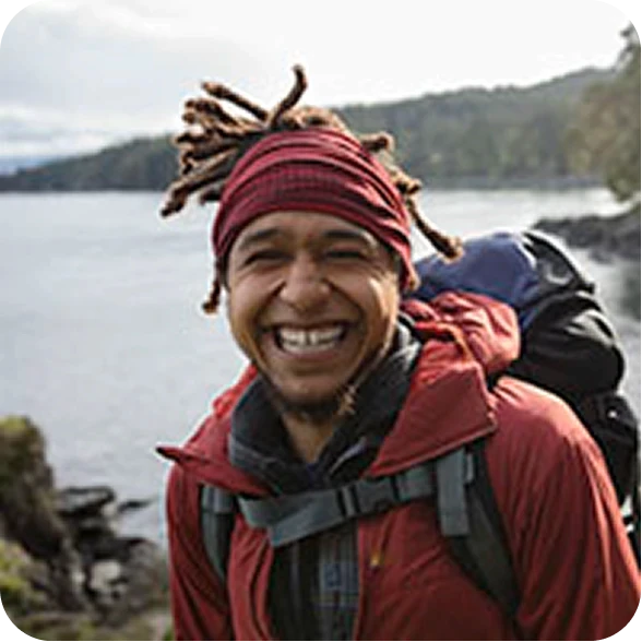 Happy man with dreadlocks wearing a red jacket and backpack hiking outdoors near a lake.
