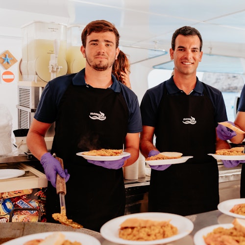 Two men in black aprons and purple gloves serving plates of food from a kitchen counter.