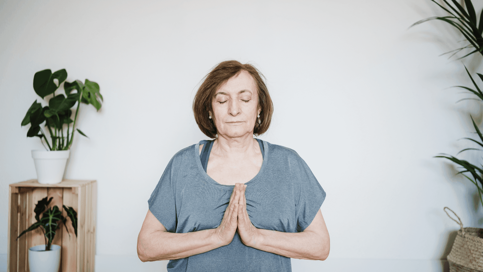 Senior woman practicing Anjali Mudra (Prayer Pose) with closed eyes, wearing grey yoga top, demonstrating mindful meditation in serene home setting with houseplants