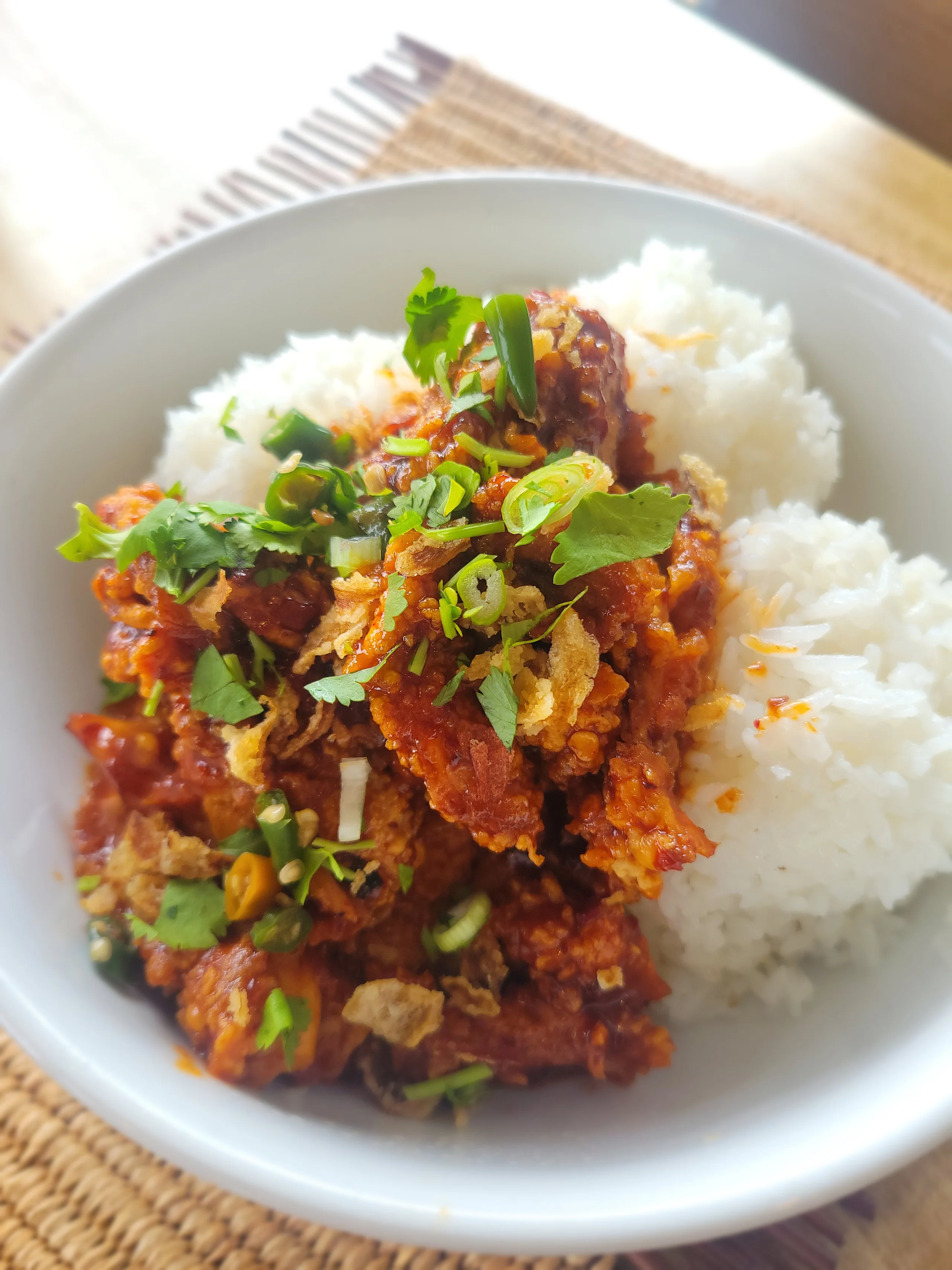 A white bowl featuring crispy fried chicken pieces coated in a red spicy sauce, garnished with cilantro, green onions, and fried shallots, served next to three scoops of white rice.