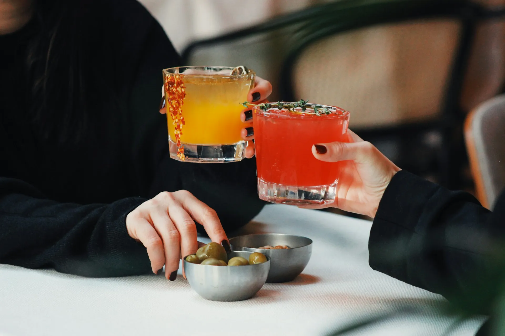 Two people clinking colorful cocktails at a bar table, with olives in small metal bowls and a casual lounge setting in the background.