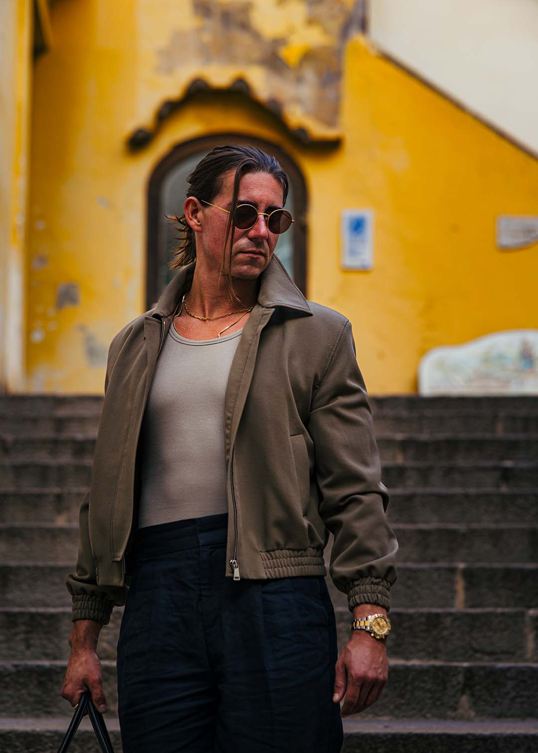 Man in a neutral bomber jacket and tailored trousers standing on stone steps in Italy, capturing modern European style and confidence.