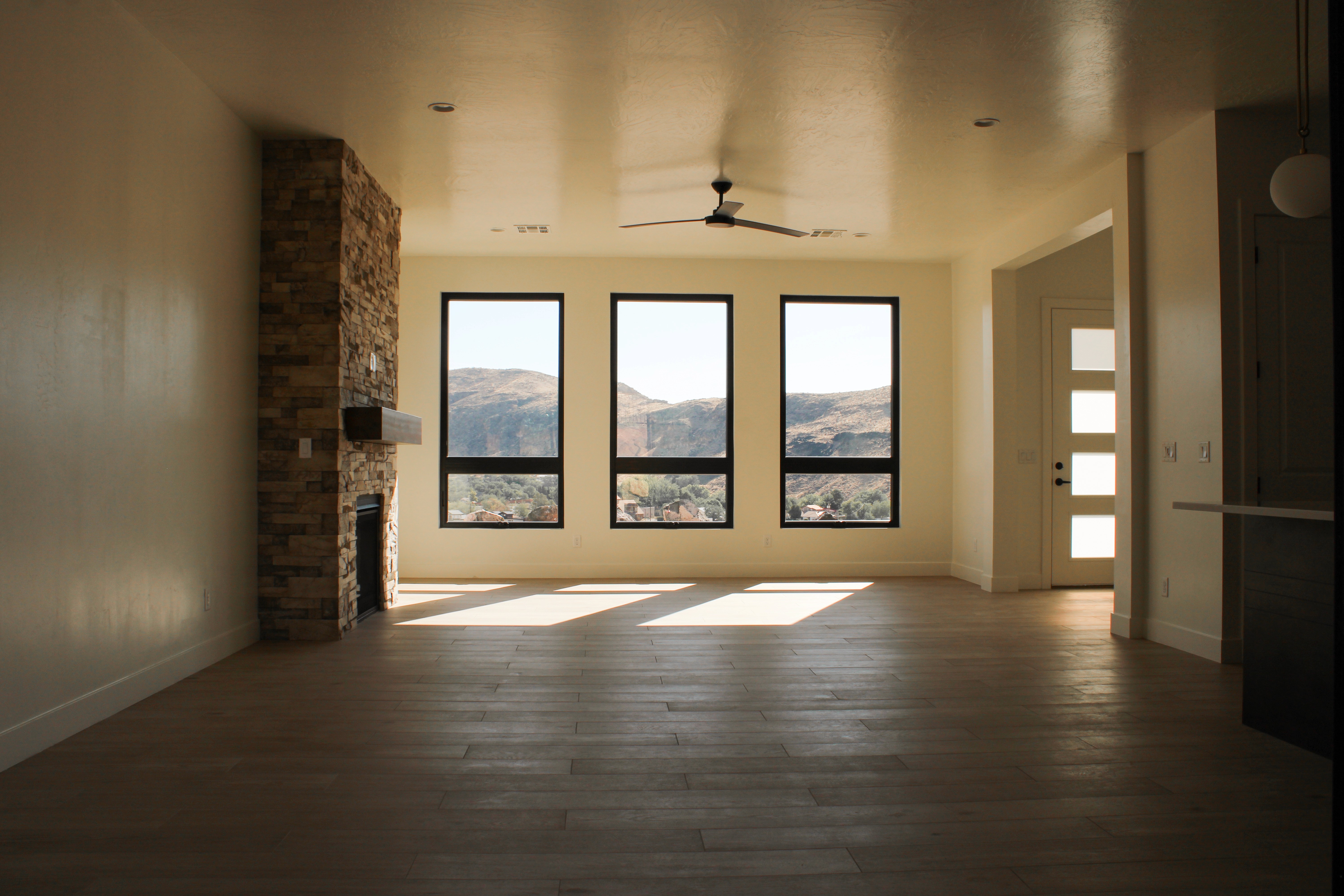 Open living room in a custom home in Hurricane, Utah featuring a stacked stone fireplace, wood-look tile floors, and large black-framed windows with mountain views.