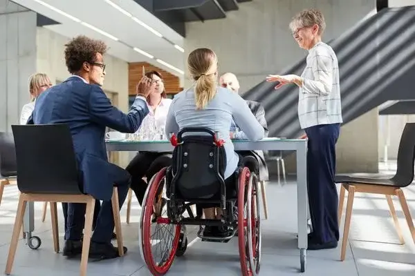 A imagem mostra um grupo de pessoas em uma reunião de negócios, todas reunidas ao redor de uma mesa de conferência. Entre elas, uma pessoa em cadeira de rodas está de costas, participando ativamente da discussão. O ambiente é moderno, com iluminação natural, e uma mulher mais velha, de pé, parece estar liderando a conversa ou fazendo uma apresentação. As outras pessoas na mesa estão engajadas, sugerindo um ambiente inclusivo e colaborativo.