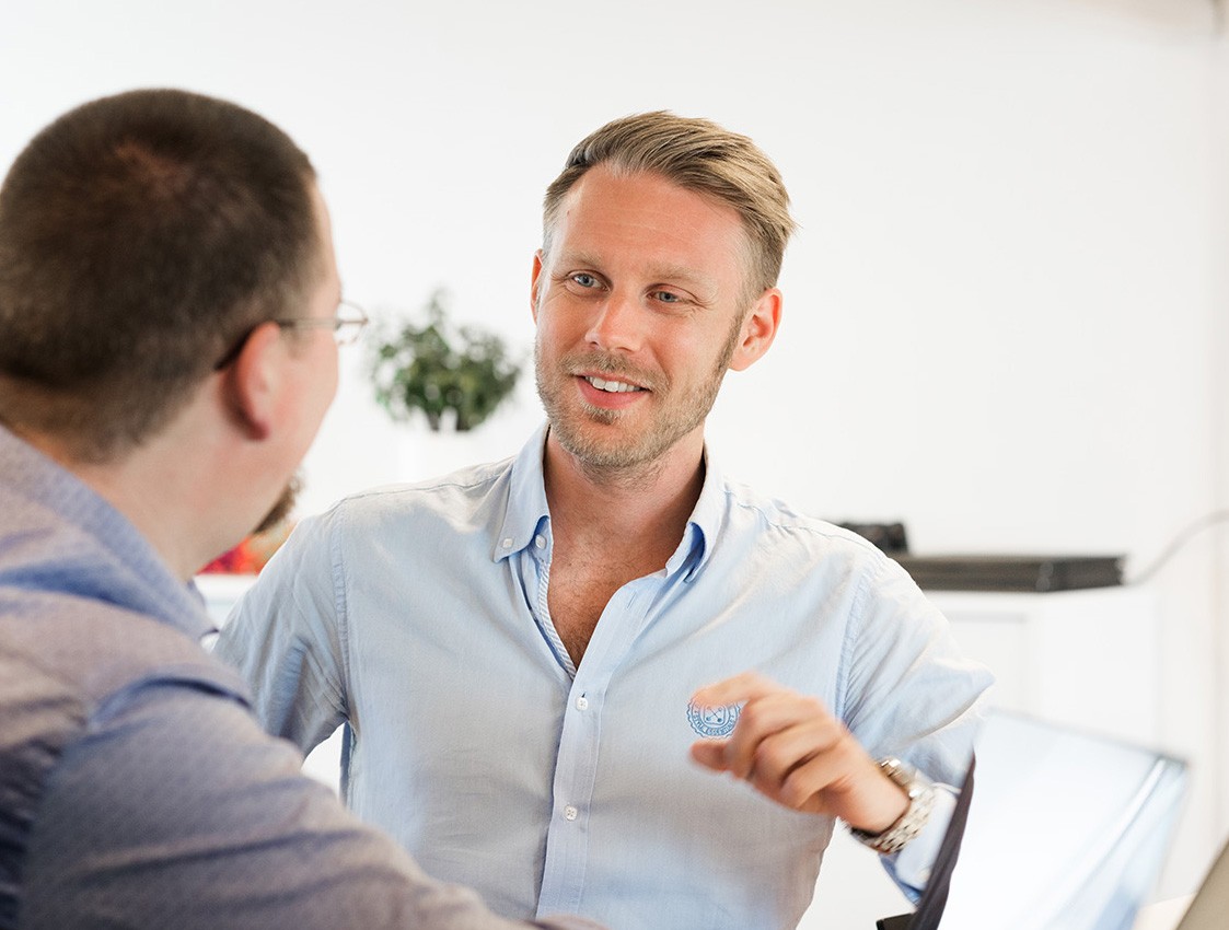 A smiling young man with crossed arms, wearing a plaid shirt and white t-shirt, poses against a dark background.